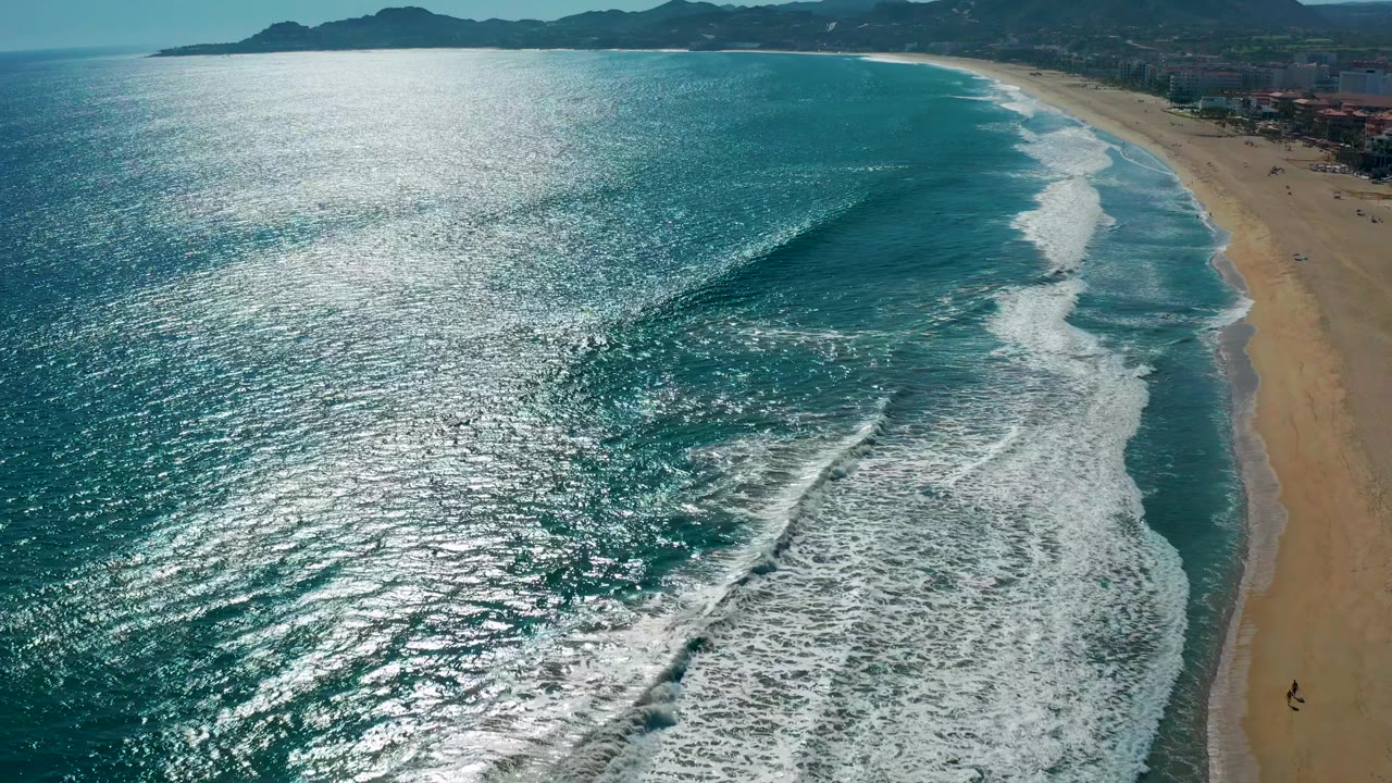 Aerial San Jose del Cabo shoreline with a large foamy break sweeping in from the lower center, bright sun reflection across the water, sandy beach at right, and mountains along the horizon.