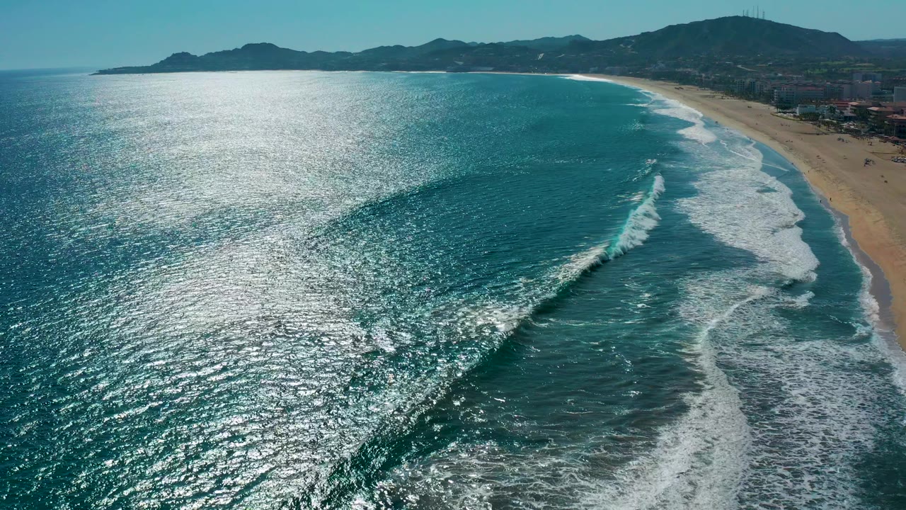High aerial frame of San Jose del Cabo shoreline where multiple white foam lines trace the beach, with sparkling turquoise water in the foreground and no visible people.