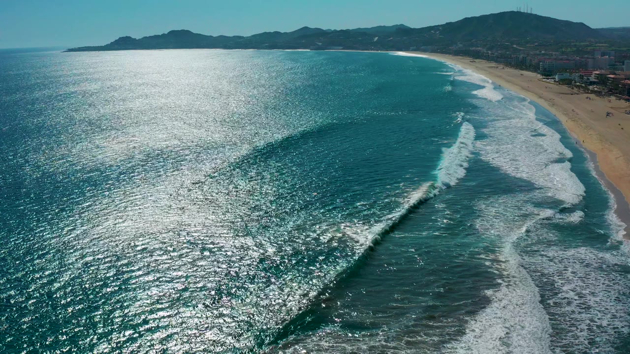 San Jose del Cabo aerial coastline with a darker wave face curling toward the shore, bright sun reflection on the water, distant mountains, and no visible people.