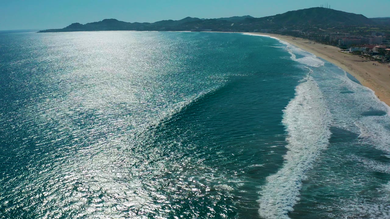 Aerial frame over San Jose del Cabo showing turquoise ocean, bright sun glitter, a white wave line nearing the sandy beach, distant hills, coastal buildings, and no visible people.