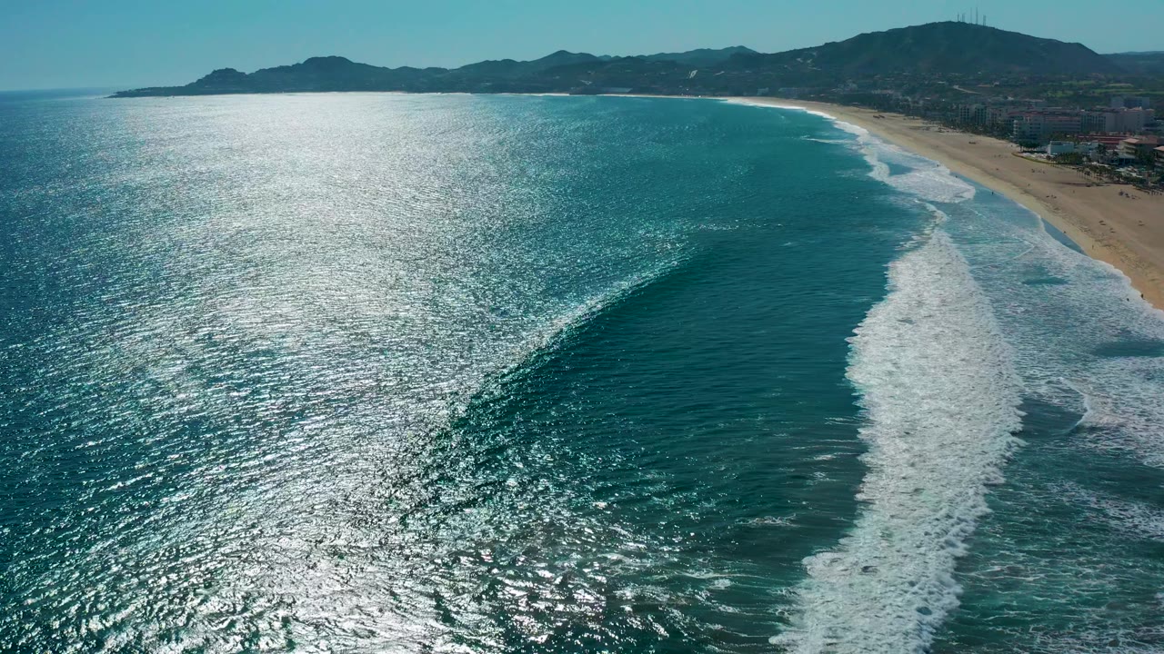 Aerial view in San Jose del Cabo with the breaking wave curling through the center-right water, sandy beach along the edge, bright sea reflection, mountain horizon, and no visible people.
