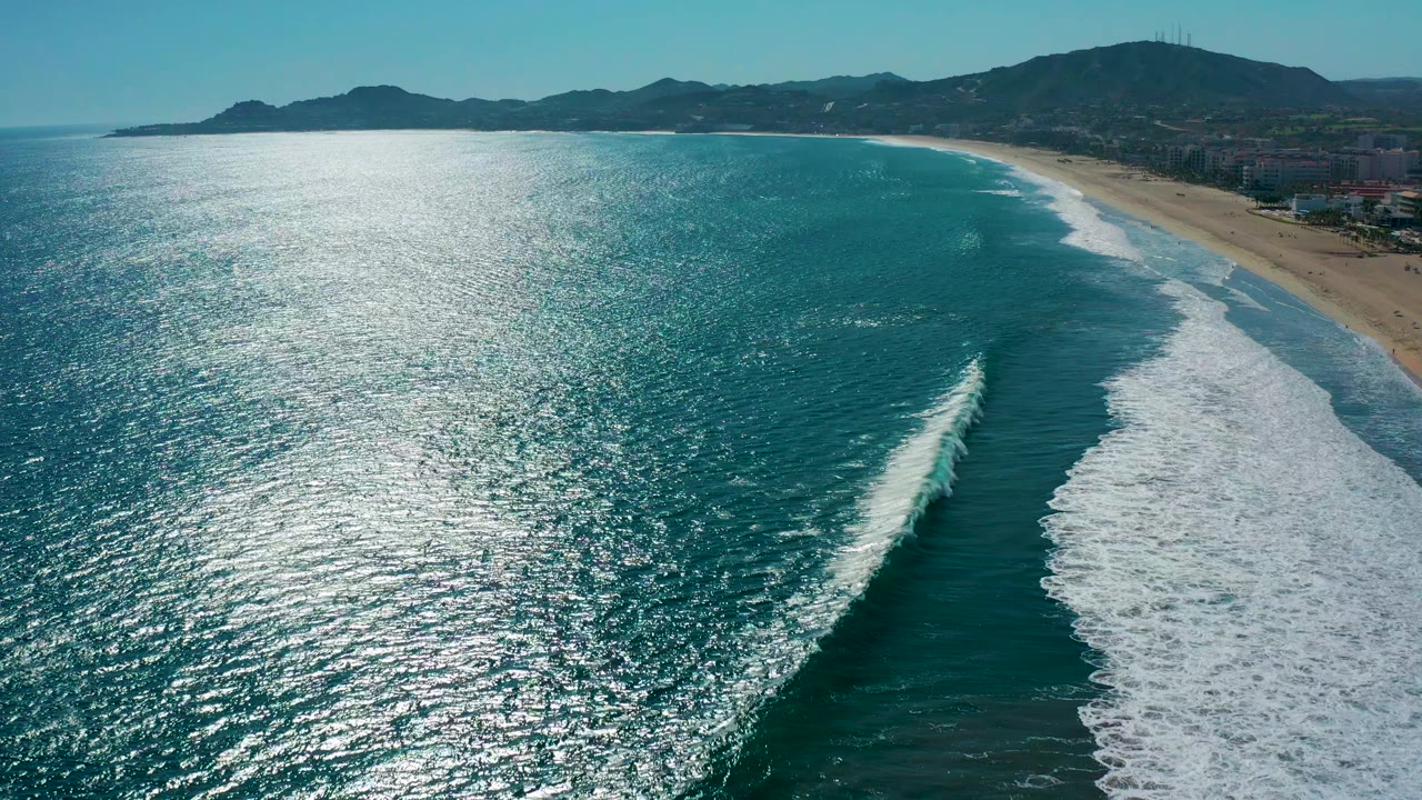 San Jose Del Cabo coastline seen from above, with a bright shimmer path on the ocean, diagonal surf lines, beach at right, hills beyond, and no visible people.
