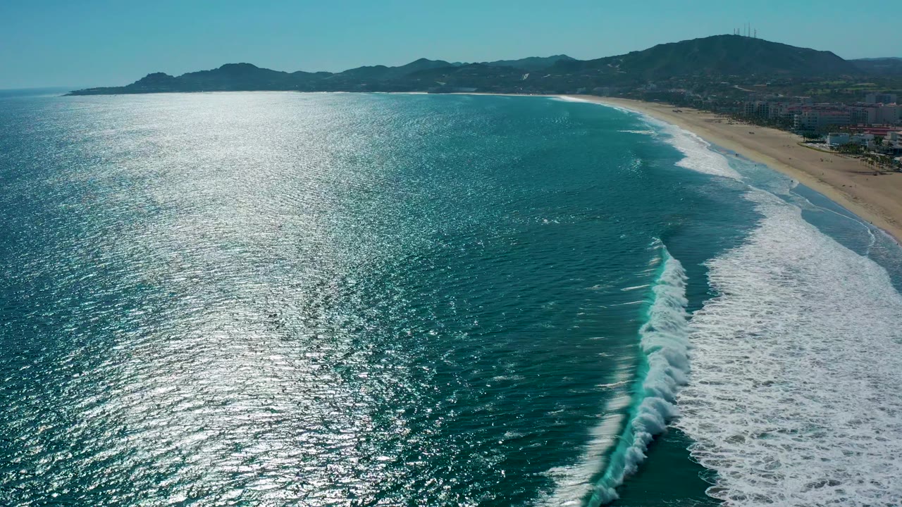 High aerial view in San Jose Del Cabo, Mexico, showing a broad sheet of shimmering ocean, surf rolling in bands, sandy beach along the right edge, and no visible people.