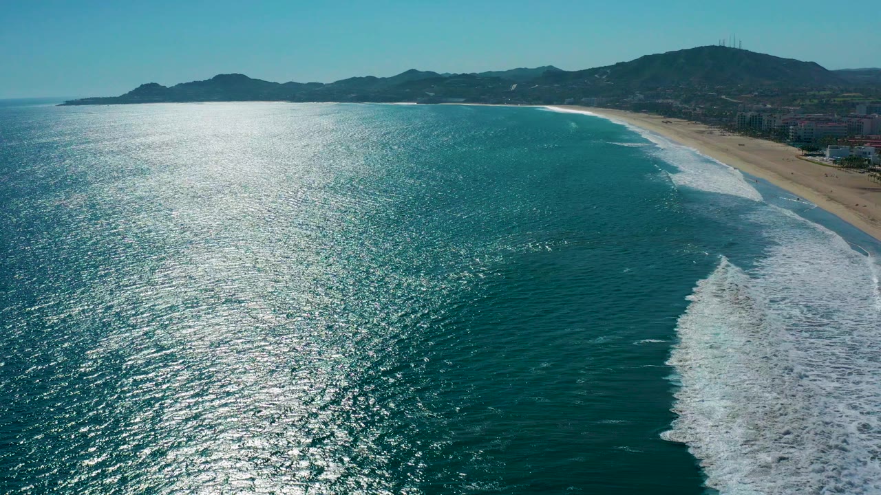 Aerial view over San Jose Del Cabo with sunlit blue ocean on the left, white surf curling toward the sandy beach on the right, mountains in the distance, and no visible people.