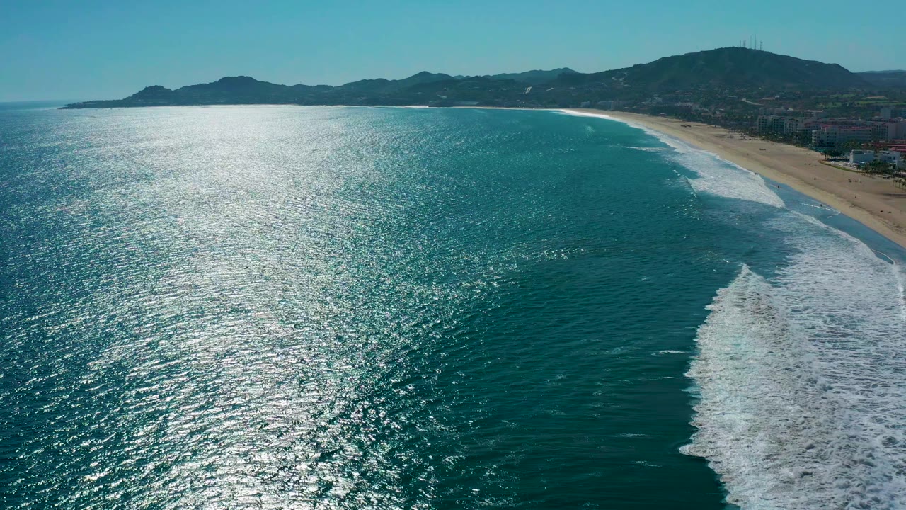 Blue coastal water with bright sun glare, breaking waves, sandy beach, and mountains.