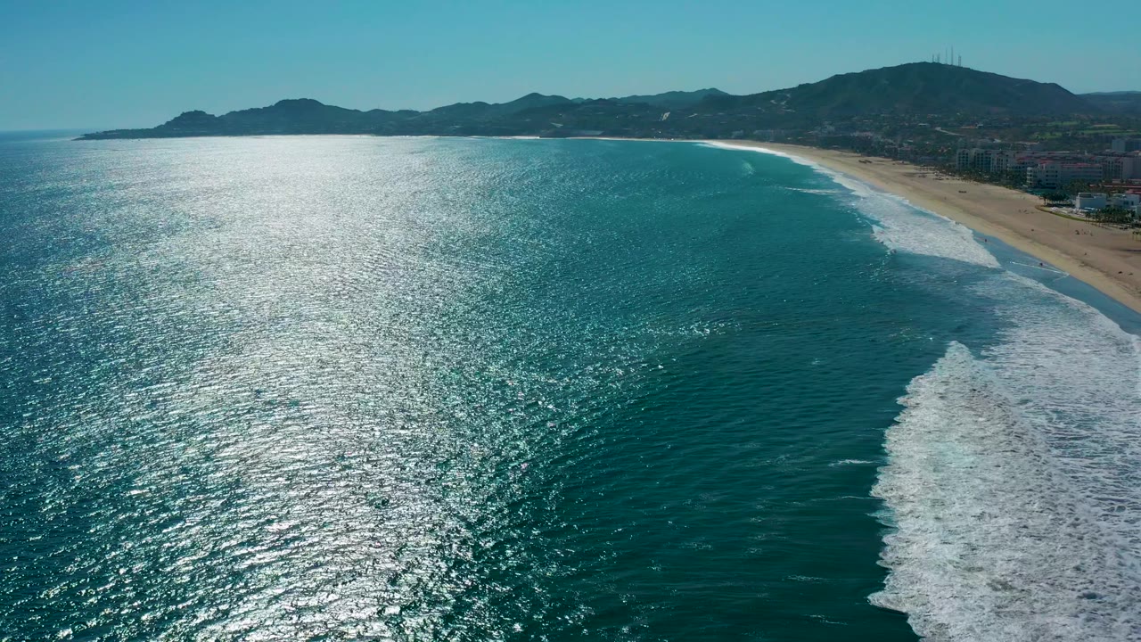 San Jose Del Cabo coastline seen from above with turquoise water filling the frame, foam gathering along the right edge, sandy beach beyond it, dark hills on the horizon, and no visible people.