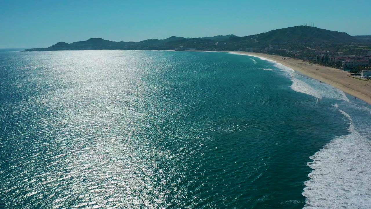 Aerial view over San Jose Del Cabo coastline with turquoise ocean in the foreground, white surf reaching the sandy beach, distant mountains, clear sky, and no visible people.