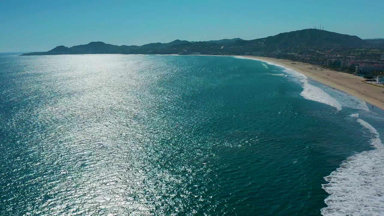 Aerial frame over the San Jose Del Cabo shore with turquoise water in the foreground, white surf rolling diagonally toward the beach, distant mountains, clear sky, and no visible people.