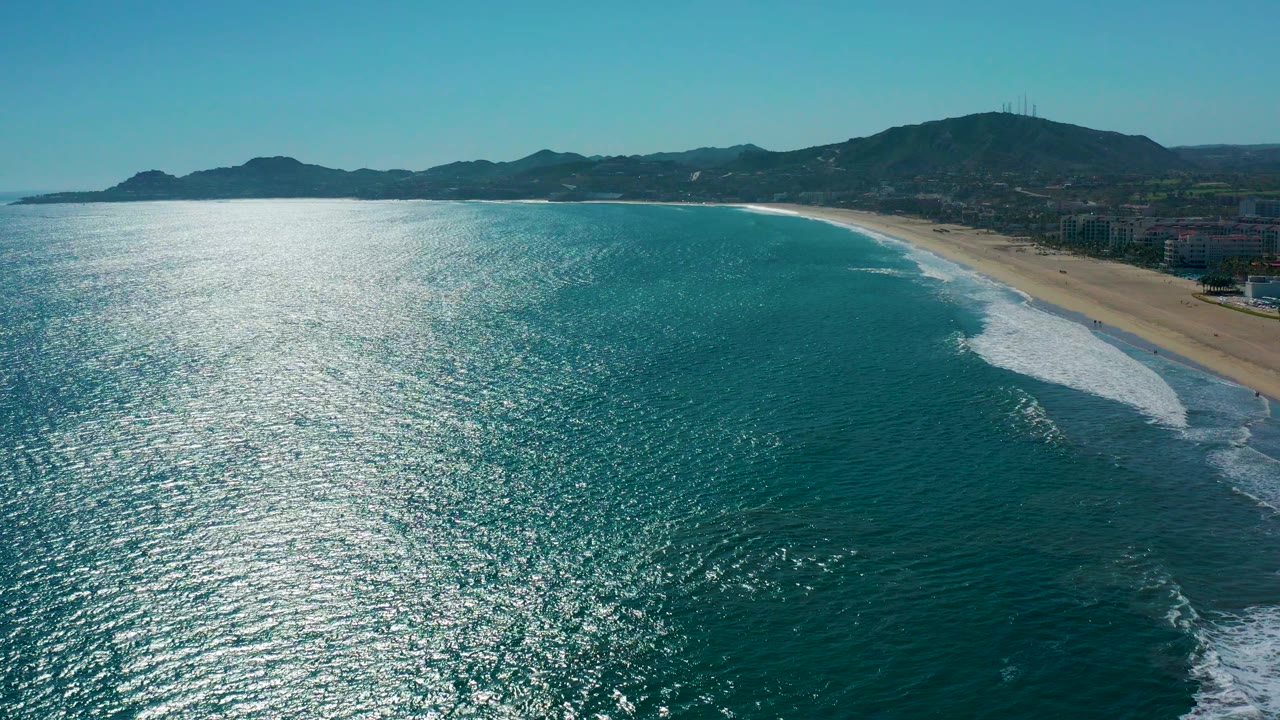 Opening aerial frame over San Jose Del Cabo, Mexico, with bright sun glare on turquoise water, white surf along the sand, distant mountains, coastal buildings, and no visible people.
