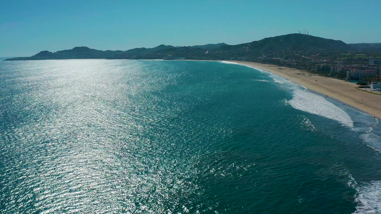 Aerial view one second in over San Jose Del Cabo, Mexico, showing a long band of shimmering ocean, a curved sandy shoreline, white breaking waves, distant hills, and no visible people.