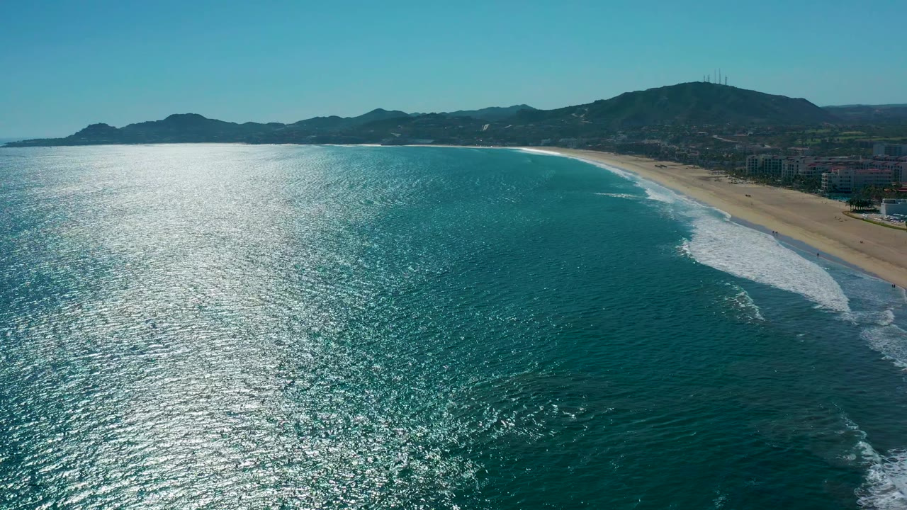 Aerial frame over San Jose Del Cabo, Mexico, with the ocean taking most of the view, white surf bands nearing the beach, mountains across the horizon, and no visible people.