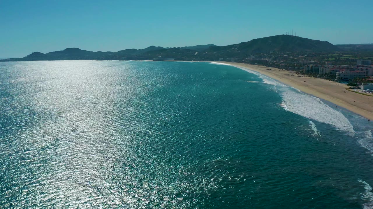 Mid-clip aerial frame over San Jose Del Cabo, Mexico, where strong sunlight sparkles on the water, waves line the sandy beach, distant mountains sit beyond low buildings, and no visible people appear.