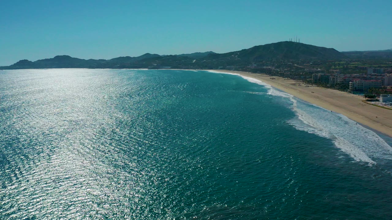 Aerial frame over San Jose del Cabo with sunlit blue-green ocean in the foreground, a wide sandy beach to the right, low hills beyond, and no visible people.