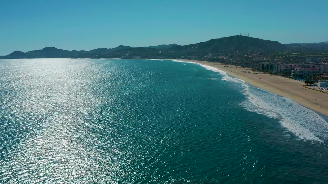Aerial San Jose del Cabo coastline with broad ocean glare across the left foreground, foamy waves meeting the beach, distant low hills, shoreline buildings, and no visible people.