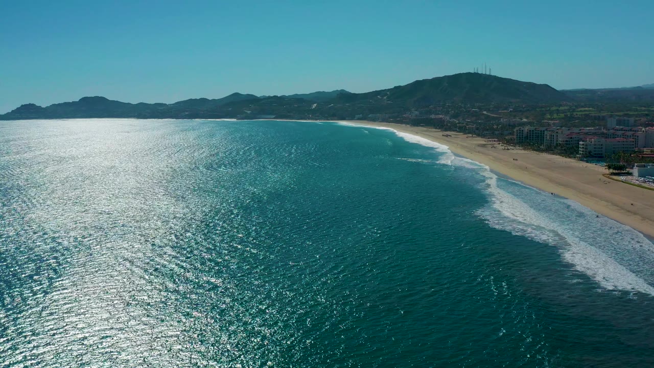 Aerial beach view with turquoise water, white surf, buildings, and hills.