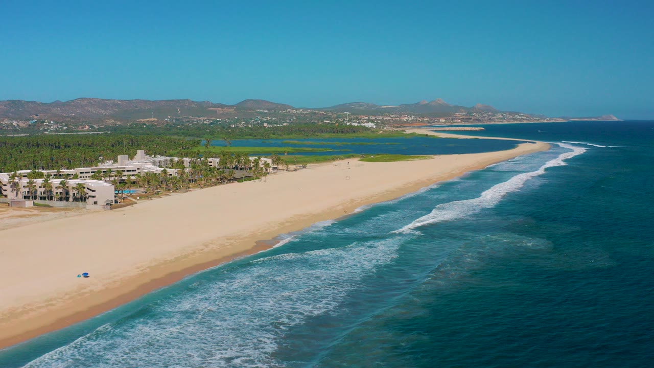 Aerial frame of San Jose del Cabo beach with pale sand at left, white surf bands over blue water at right, coastal buildings and palms inland, lagoon greenery beyond, and no visible people.
