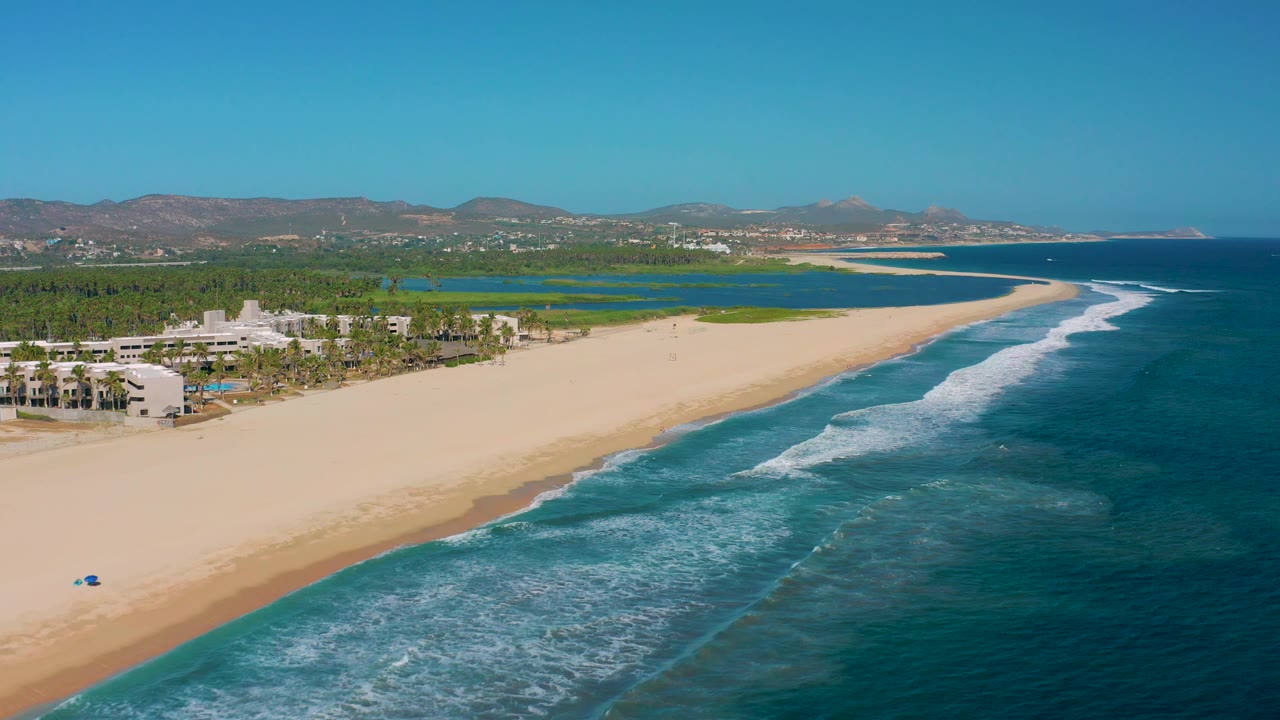 High aerial view over beach and ocean with buildings at the left edge, lagoon greenery, and distant mountains.