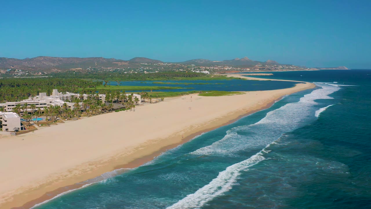 High aerial frame of San Jose Del Cabo coastline with white wave lines along the beach, pale buildings and palms at left, green wetland water behind the sand, distant hills, and no visible people.
