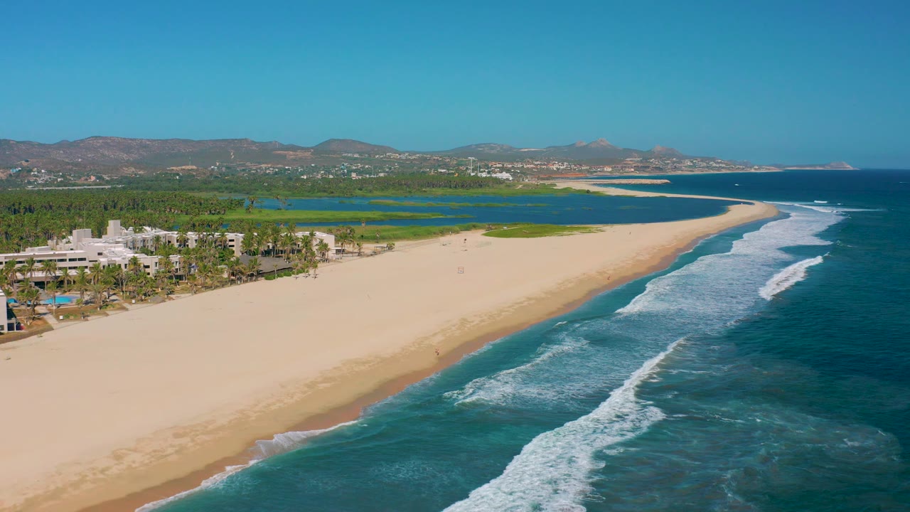 San Jose Del Cabo aerial frame with resort buildings partly cropped at the left edge, a wide empty-looking sand area, white surf bands on the right, green wetlands beyond the beach, and no visible people.