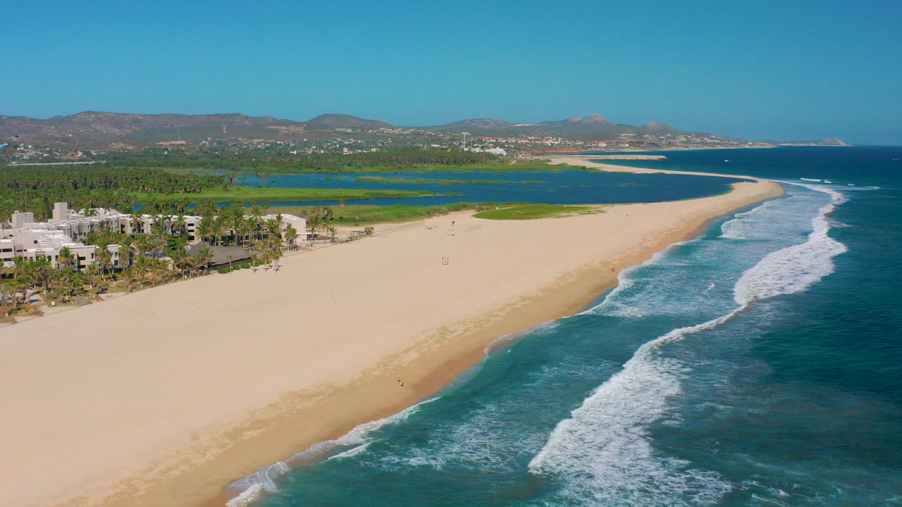 Opening aerial frame of San Jose del Cabo showing a broad sandy beach, blue surf at right, green lagoon behind the shore, palms and low buildings at left, and no visible people.
