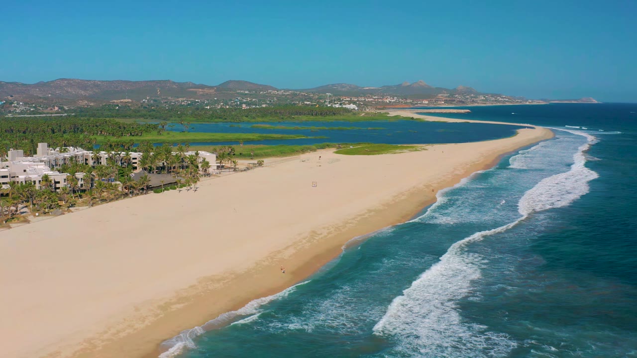 Early aerial view of San Jose del Cabo with the beach stretching farther across the foreground, waves breaking along the right edge, lagoon water behind the sand, and no visible people.