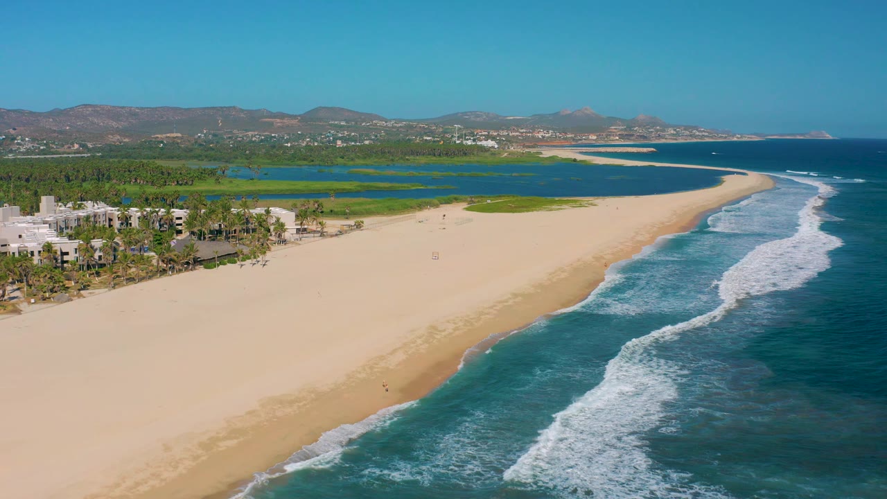 Aerial frame over San Jose del Cabo where the right-side surf advances in white bands, the open sand dominates the center, and the lagoon, palms, and low buildings sit behind it with no visible people.