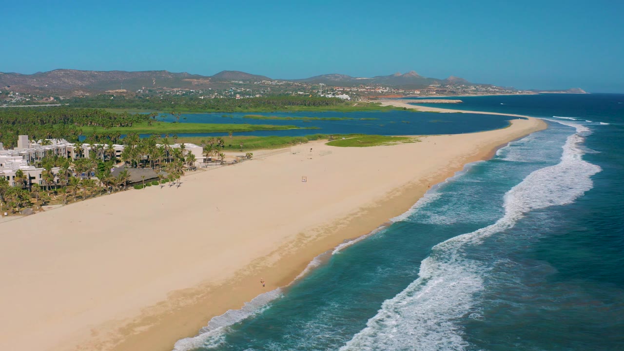 Diagonal shoreline with surf, pale sand, lagoon, palms, buildings, and mountain backdrop.