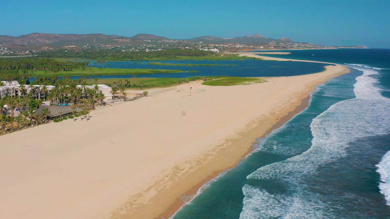 San Jose Del Cabo aerial frame with pale sand curving beside blue ocean, white wave foam along the shore, lagoon water behind the beach, palms at left, hills on the horizon, and no visible people.