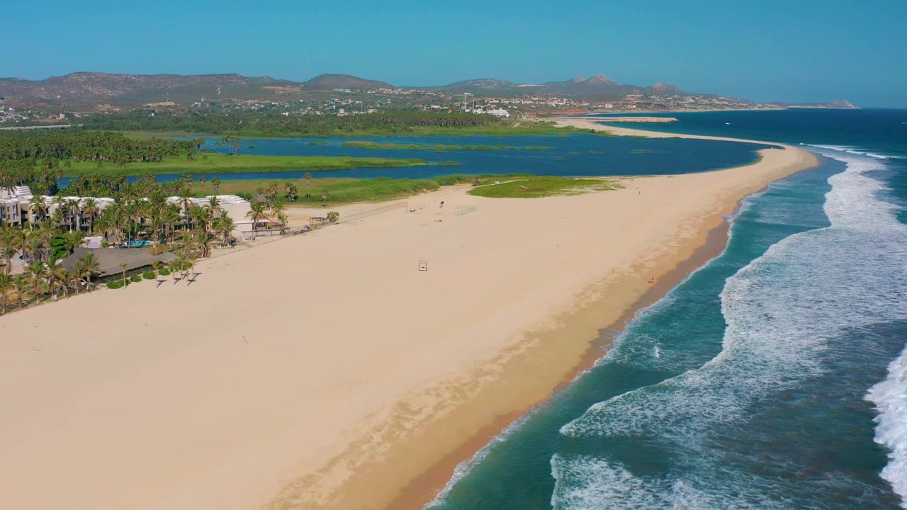 Drone-style aerial view in San Jose Del Cabo, Mexico with the shoreline running diagonally, surf breaking at right, lagoon and greenery across the background, palms near the lower left, and no visible people.