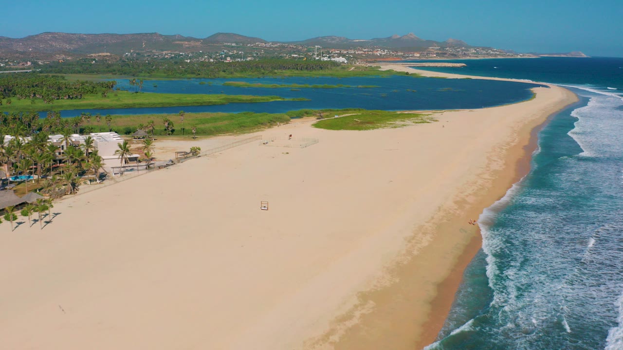 Aerial view over San Jose Del Cabo showing a wide sandy beach, blue ocean surf on the right, green lagoon inland, palms at left, distant hills, and no visible people.