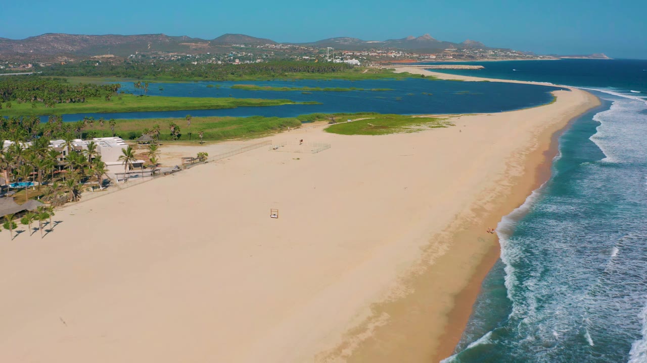 Wide aerial coastline with sandbar, lagoon, waves, palms, and hills.