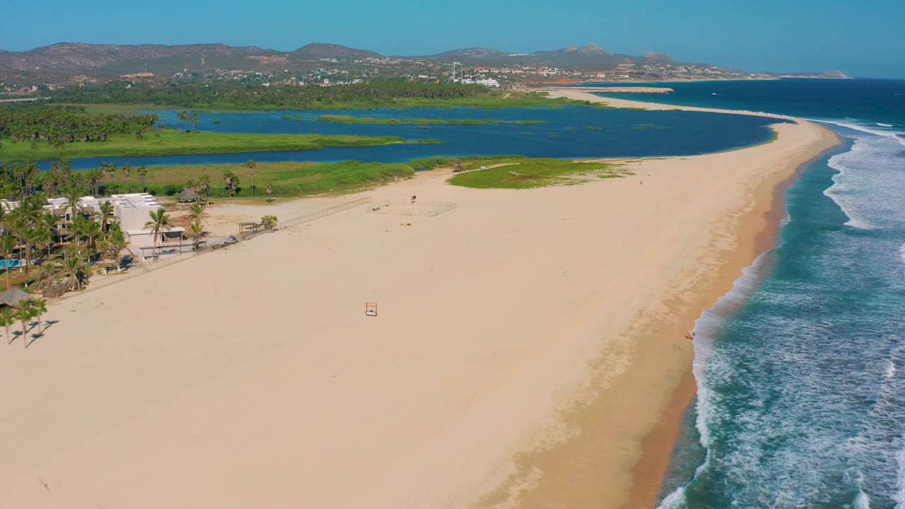 Aerial San Jose Del Cabo beach view with more open sand in the foreground, foamy ocean water on the right, the lagoon and green vegetation behind the beach, distant hills, and no visible people.