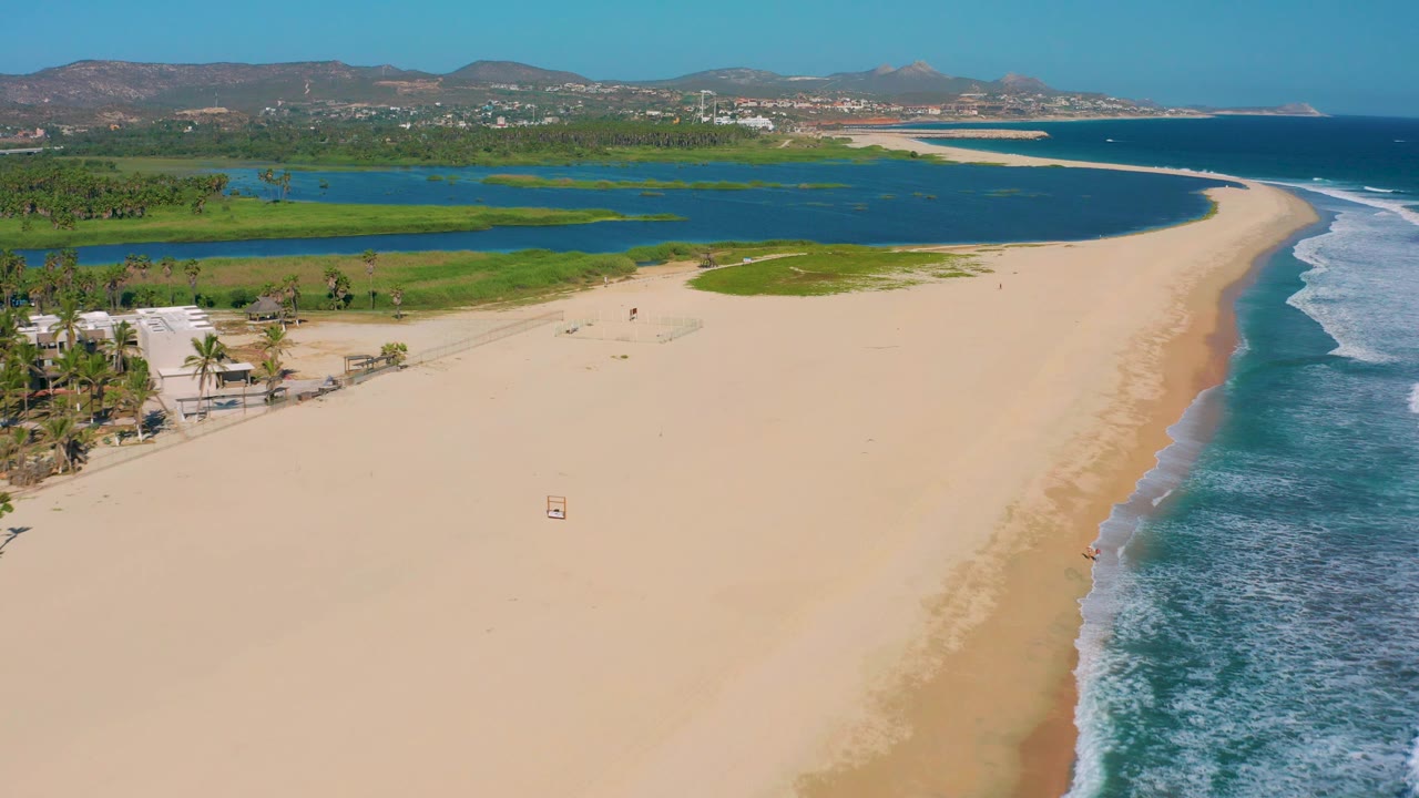 Drone view of beach, lagoon, palms, ocean waves, and hillside background.