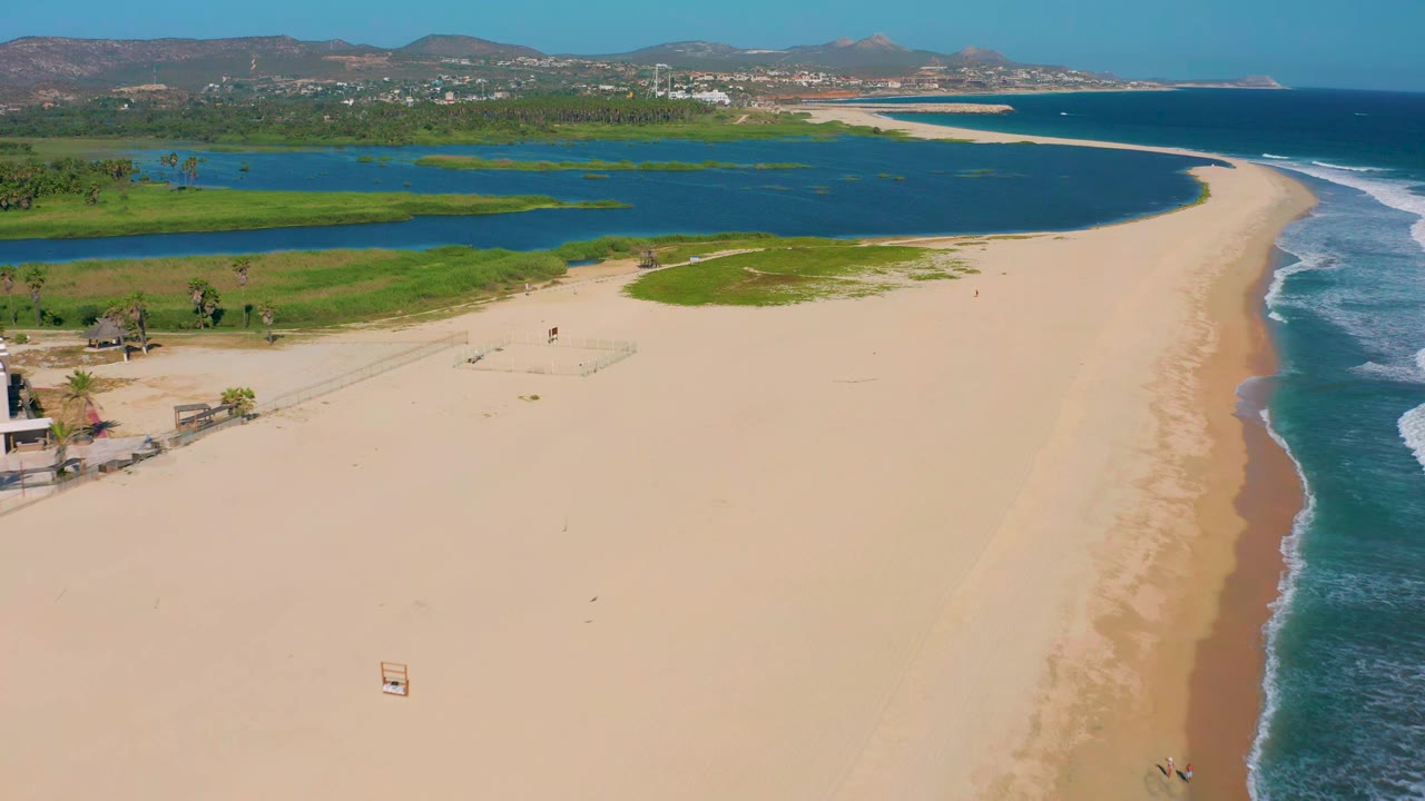 High aerial frame over San Jose del Cabo with pale sand in the foreground, calm lagoon water behind it, breaking surf along the right side, and no visible people.