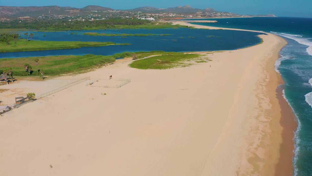 Wide aerial frame in San Jose del Cabo with open sand across the lower frame, green lagoon margins at left, ocean surf to the right, distant development, and no visible people.
