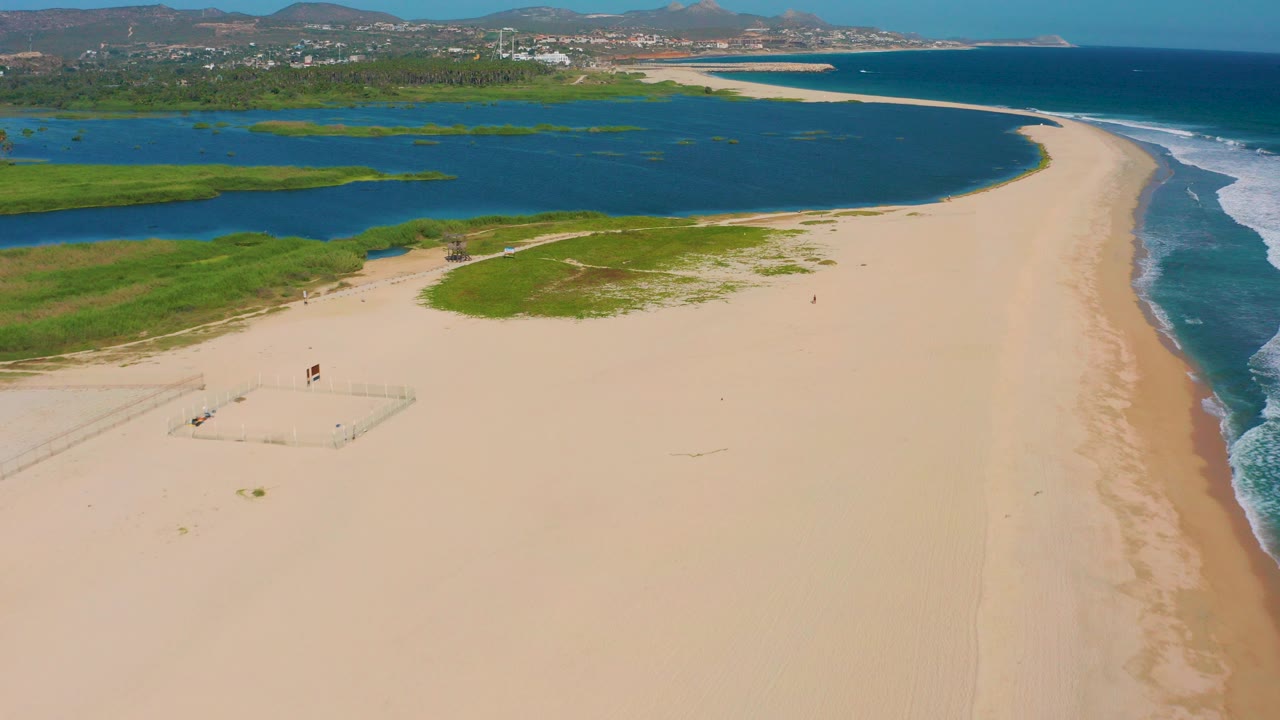 Aerial view over San Jose del Cabo showing a wide sandy beach, blue lagoon water on the left, ocean surf on the right, green vegetation, distant hills, and tiny people on the sand.