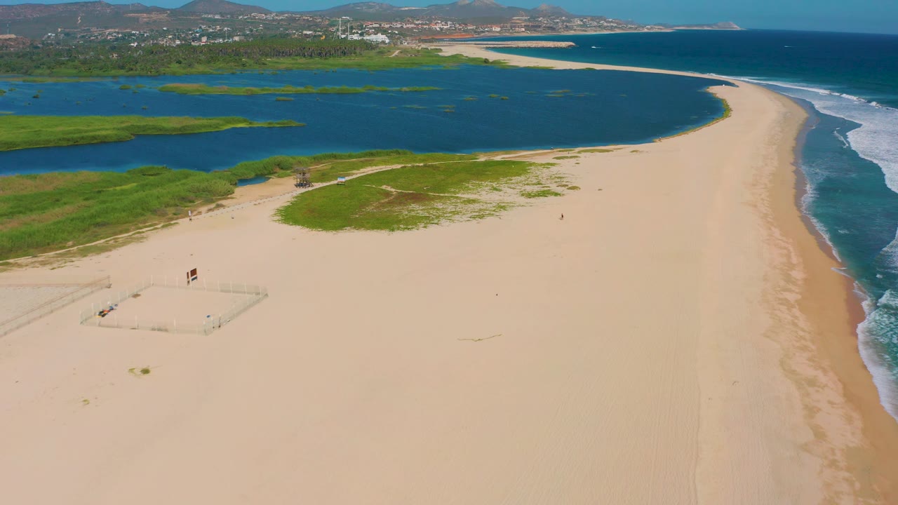 Aerial frame of San Jose del Cabo coastline with pale sand, a curved blue lagoon, white waves breaking along the ocean side, green patches near the water, and tiny beachgoers visible.