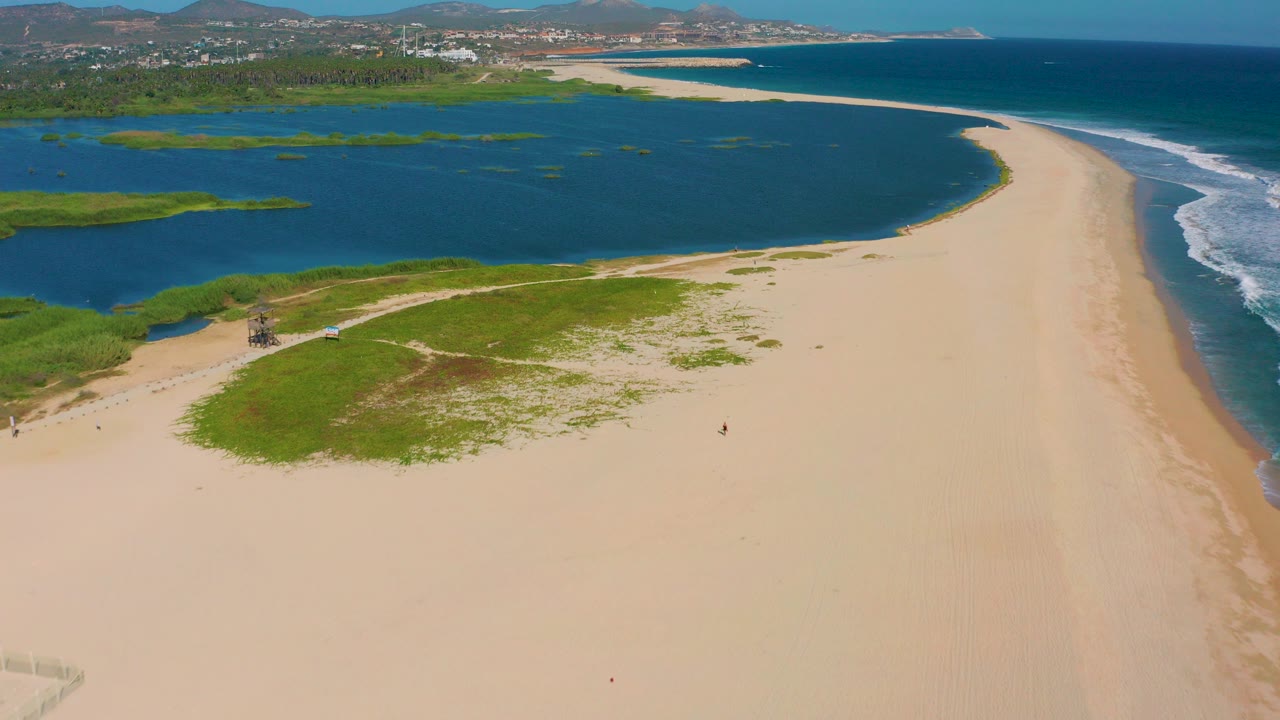 Aerial frame from San Jose Del Cabo showing a broad sandy beach below, a curved lagoon at left, ocean surf at right, green dune patches, distant shoreline buildings, and tiny people on the sand.