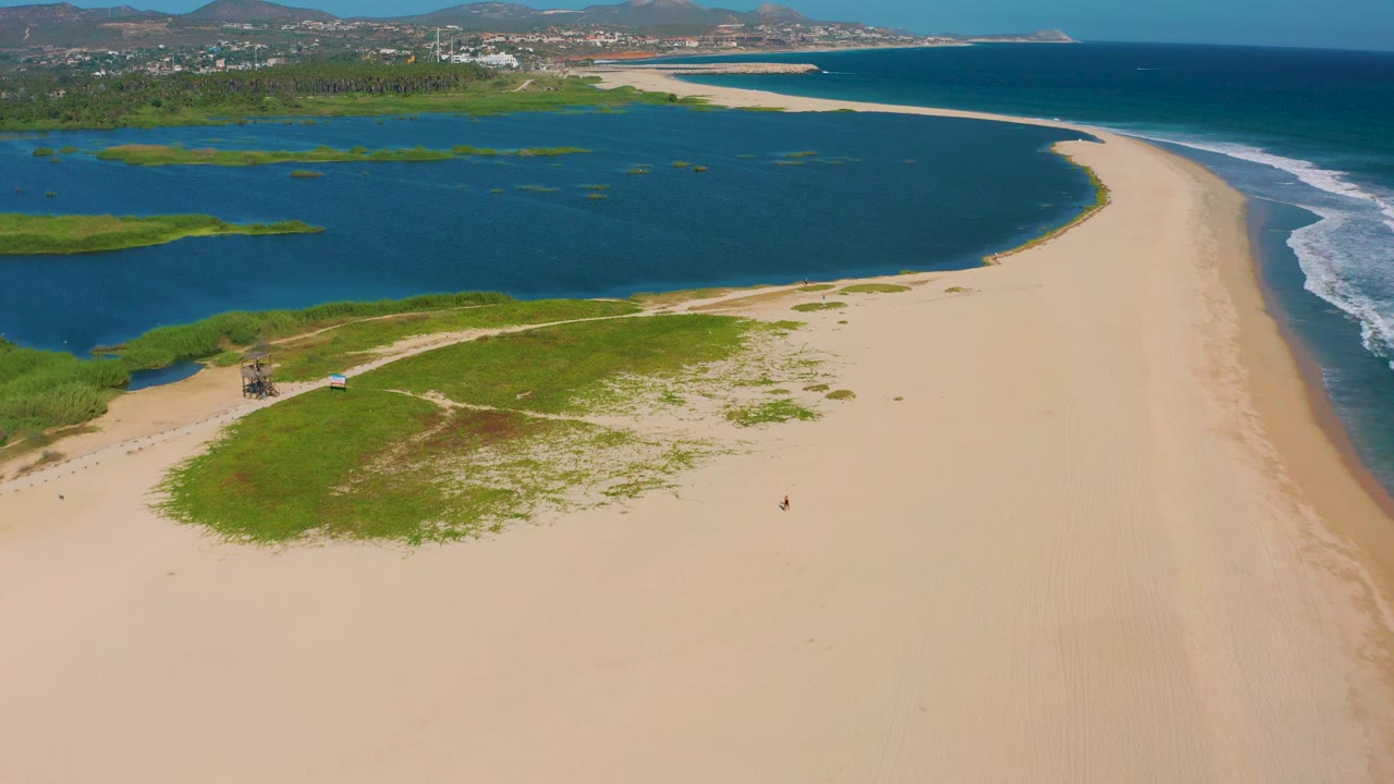 High aerial frame over San Jose Del Cabo with the pale sandbar stretching toward the background, lagoon water at left, breaking ocean waves at right, green patches near the lower edge, and distant buildings along the coast.