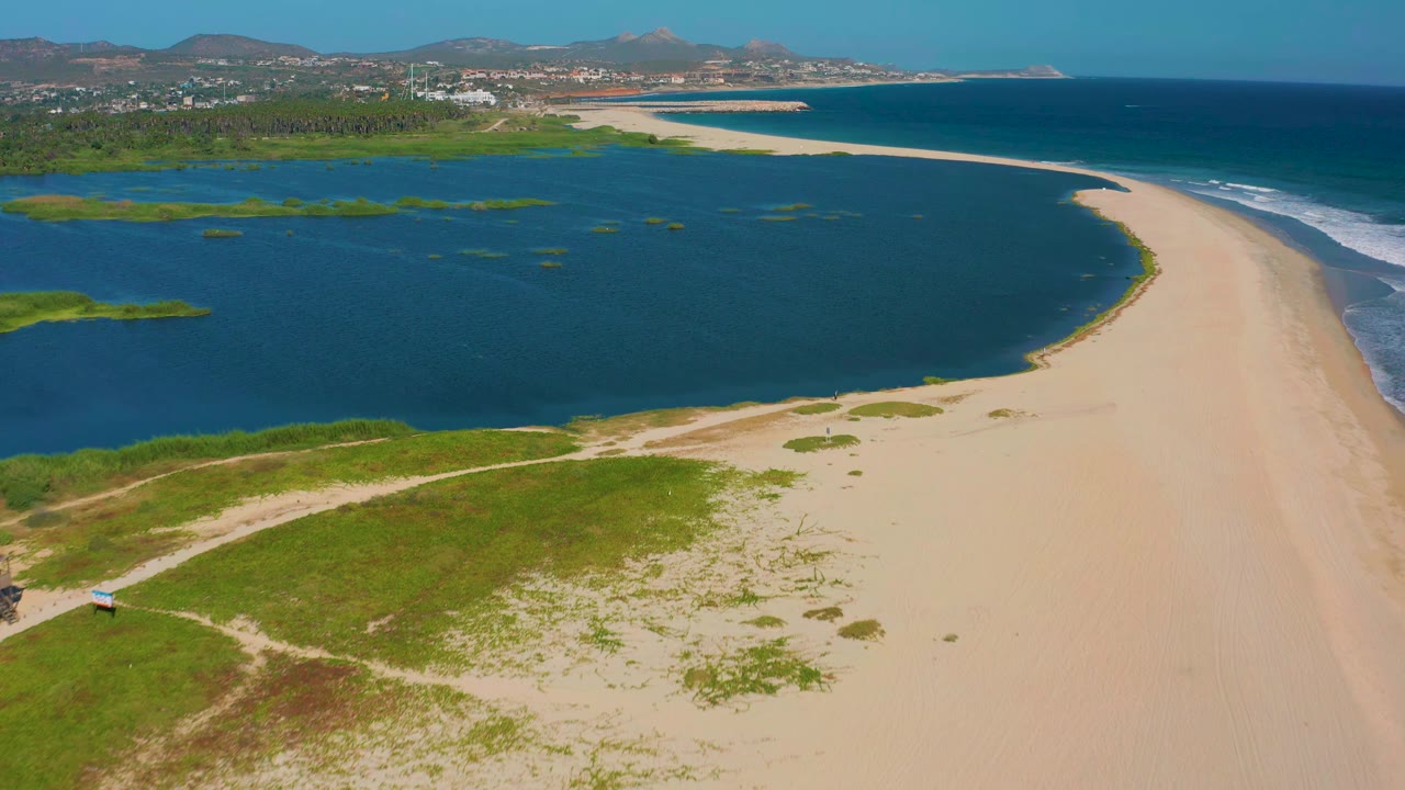 Wide aerial frame of San Jose del Cabo coastline with lagoon water on the left, a pale sandy beach curving toward the ocean, grassy foreground, distant hills, and no visible people.
