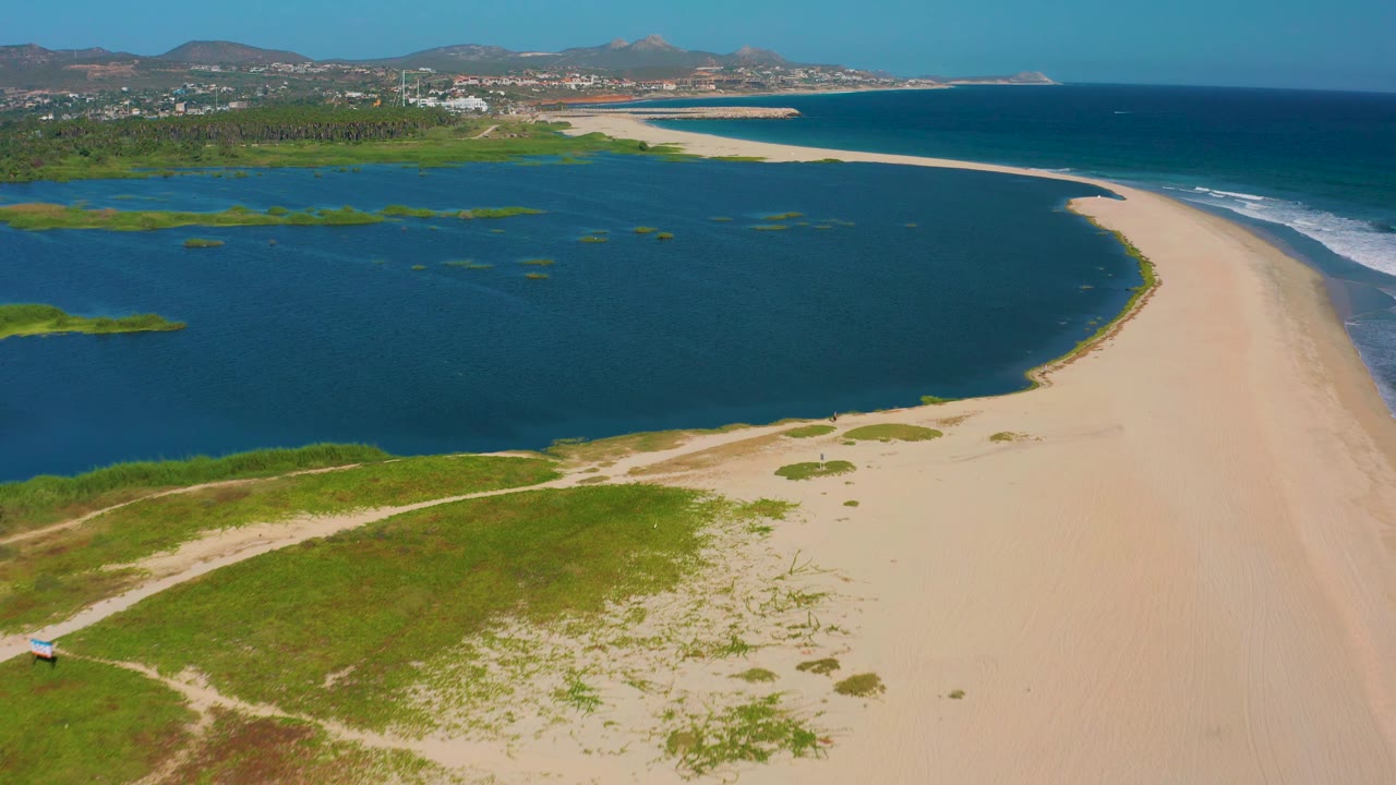Elevated San Jose del Cabo coastline view with the lagoon spreading across the left side, the beach sweeping along the right, blue ocean beyond, distant hills, and no visible people.