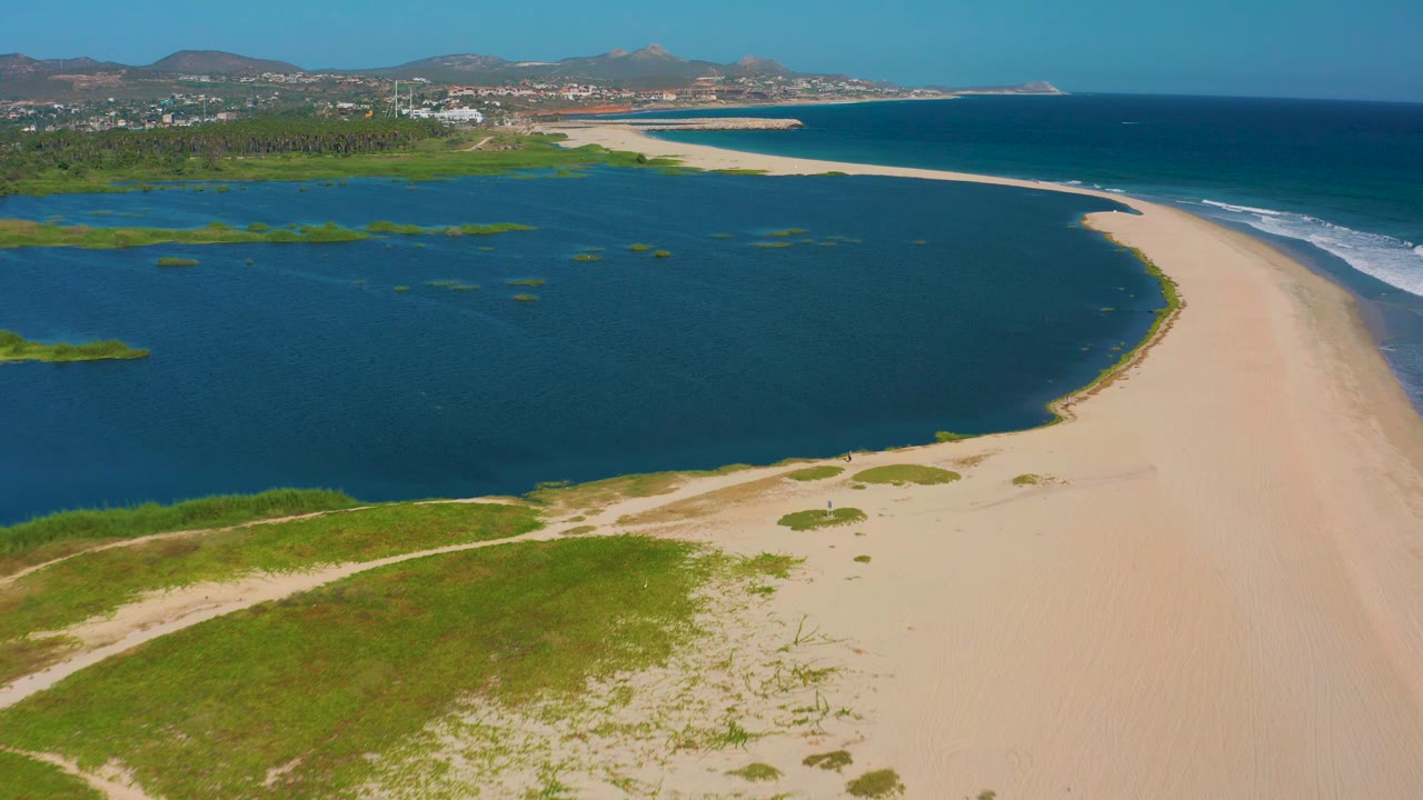 Aerial frame from San Jose del Cabo showing darker lagoon water on the left, a long empty sand strip on the right, green vegetation below, white waves offshore, and no visible people.