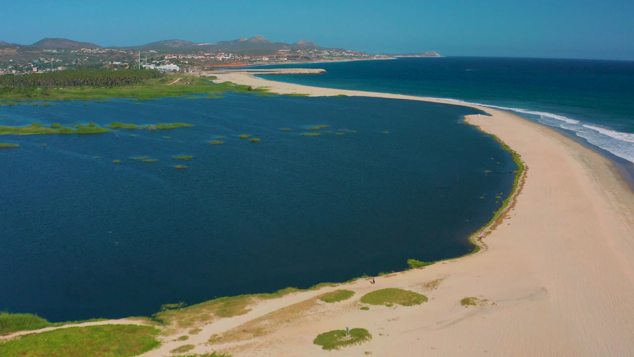 San Jose del Cabo aerial frame with dark blue lagoon in the foreground, a pale sand spit curving toward the ocean, surf on the right, distant buildings, hills, and no recognizable people.