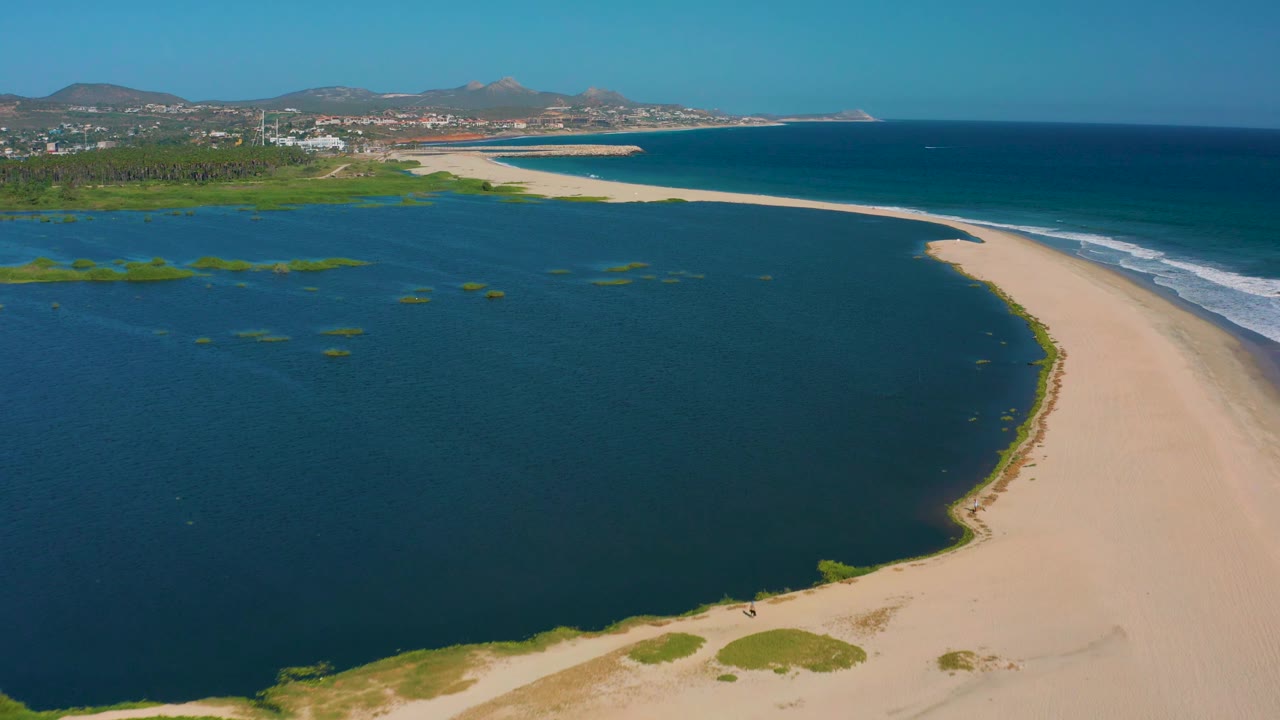 San Jose del Cabo aerial view with the lagoon taking more of the frame, the sandy beach curving along the right edge, ocean surf beyond it, and distant shoreline buildings under clear sky.