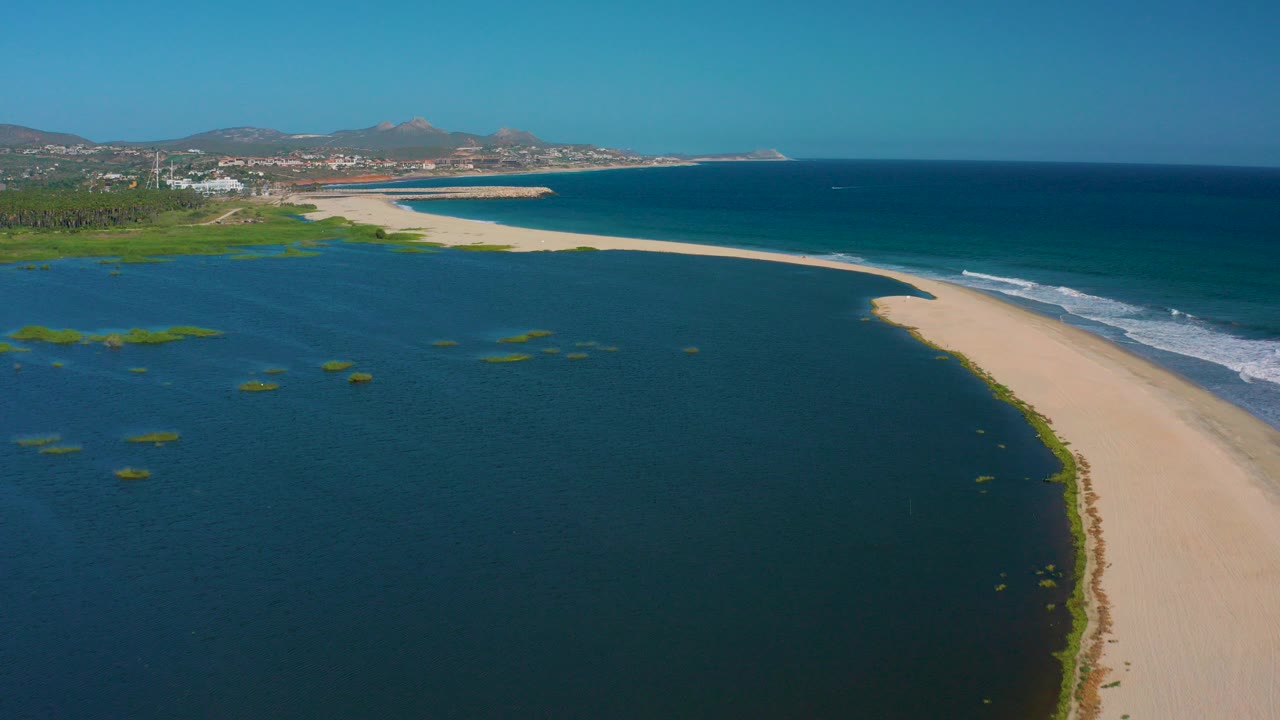 Aerial frame over San Jose del Cabo showing a pale sandbar between dark lagoon water and blue ocean surf, with distant hills and no visible people.
