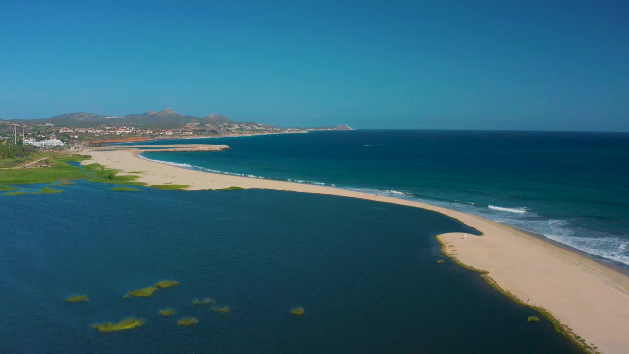 Second aerial frame over San Jose del Cabo shows the estuary filling the lower frame, green patches near the water surface, the sandbar crossing the middle, blue ocean beyond, and no visible people.