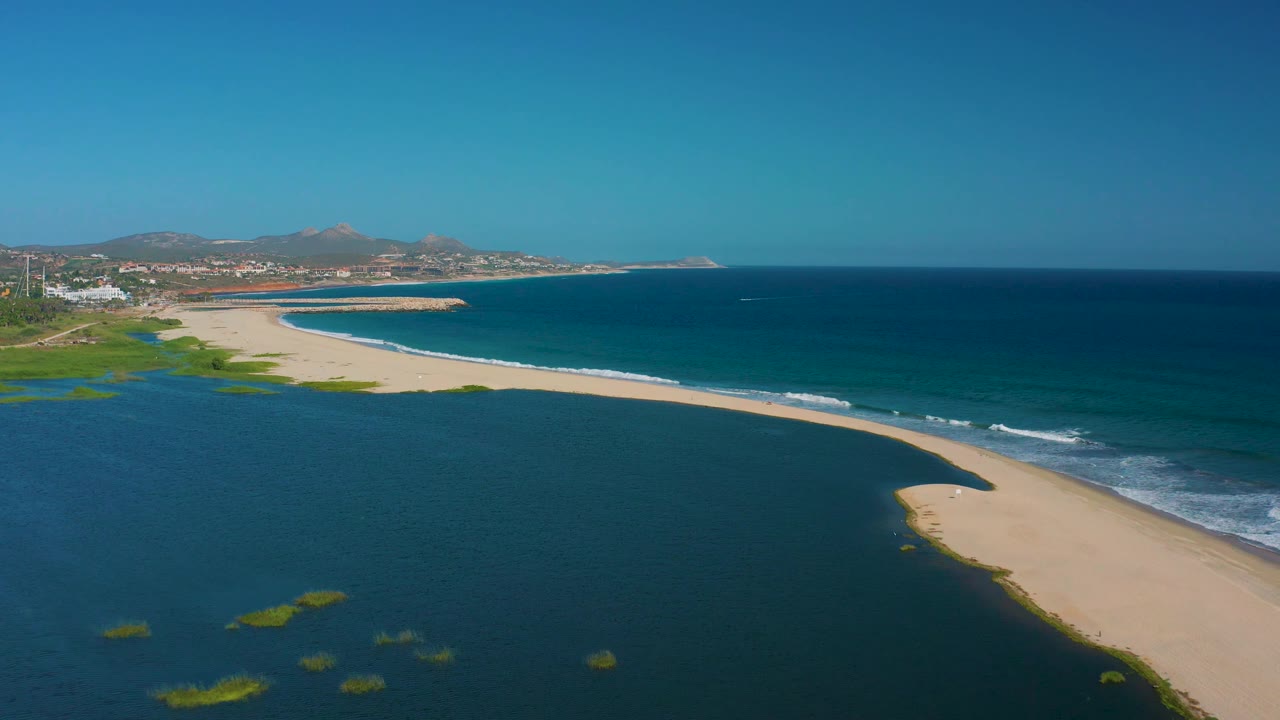 Third frame from the San Jose del Cabo aerial places more open ocean on the right, with a narrow surf line beside the sand spit, estuary water to the left, distant hills, and no visible people.