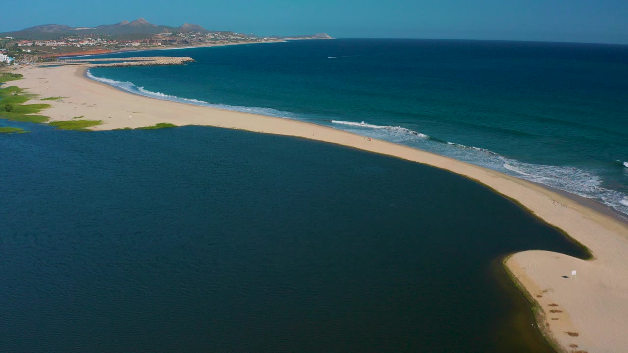 Aerial view in San Jose del Cabo, Mexico, with a pale sandbar separating a dark lagoon from blue ocean surf, distant buildings and low mountains visible beyond the coast.