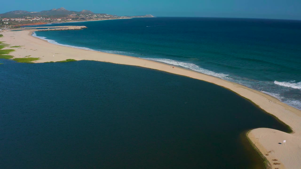 Wide aerial frame in San Jose del Cabo, Mexico, with lagoon water filling the foreground, a curved sandy beach through the center, ocean waves beyond it, and no visible people.