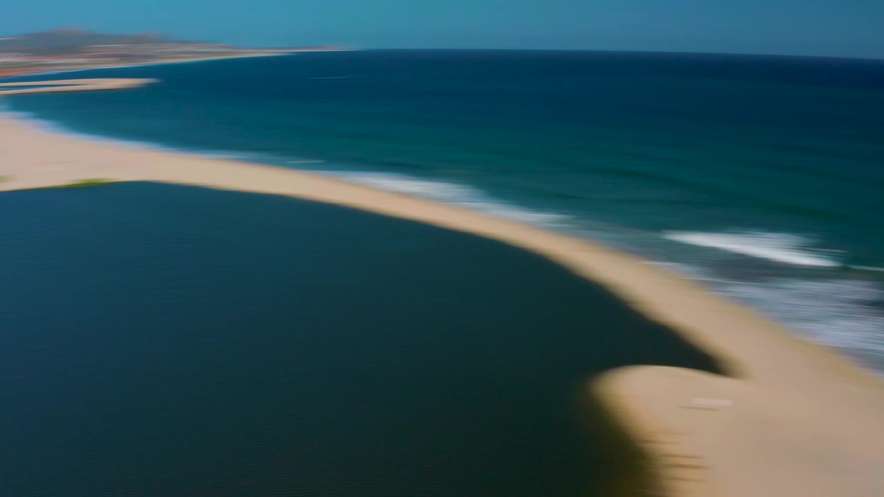 Motion-blurred aerial frame in San Jose del Cabo, Mexico, where the sandbar, lagoon, and ocean surf streak across the view, with no visible people discernible.